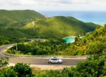 Voiture de location blanche sur une route sinueuse de Martinique, entre collines verdoyantes et mer turquoise