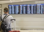 Voyageur avec un bagage cabine devant un tableau de départs dans un aéroport européen