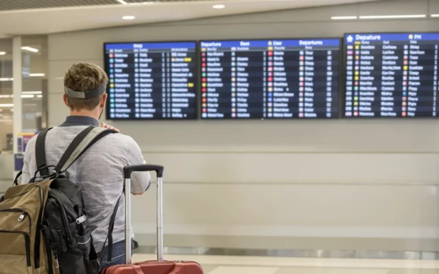 Voyageur avec un bagage cabine devant un tableau de départs dans un aéroport européen
